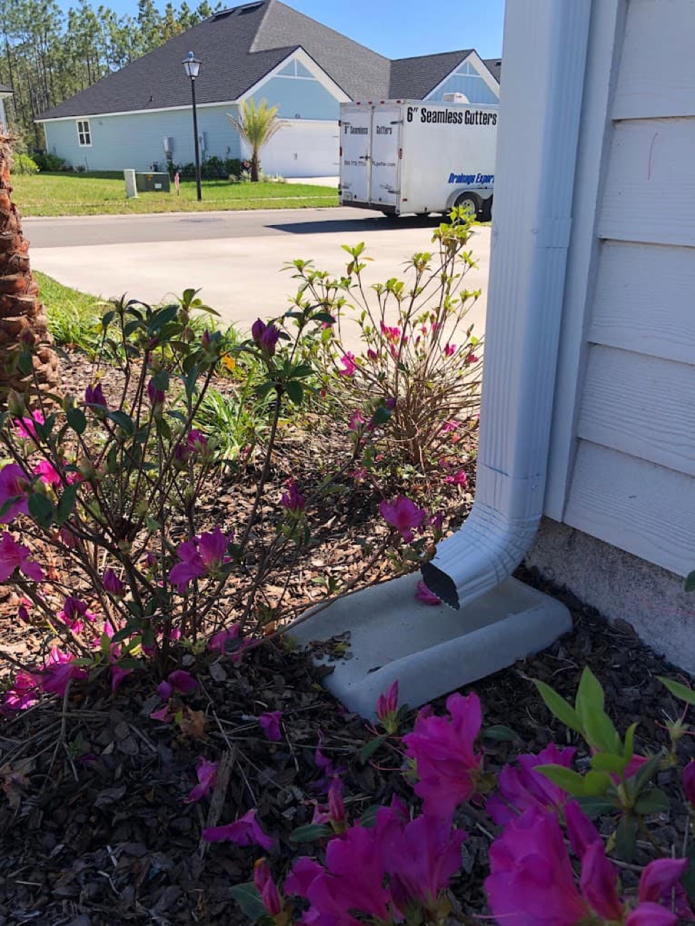 Aqua Premier 6-inch seamless gutter downspout draining onto a concrete splash block beside pink azaleas, carrying rainwater away from the home's foundation — seamless rain gutter installation across Ponte Vedra Beach, Nocatee, St. Johns, Duval & Clay County (Orange Park, Fleming Island), with the Aqua Premier Seamless Gutters & Drainage Experts trailer on site (904-779-3985)